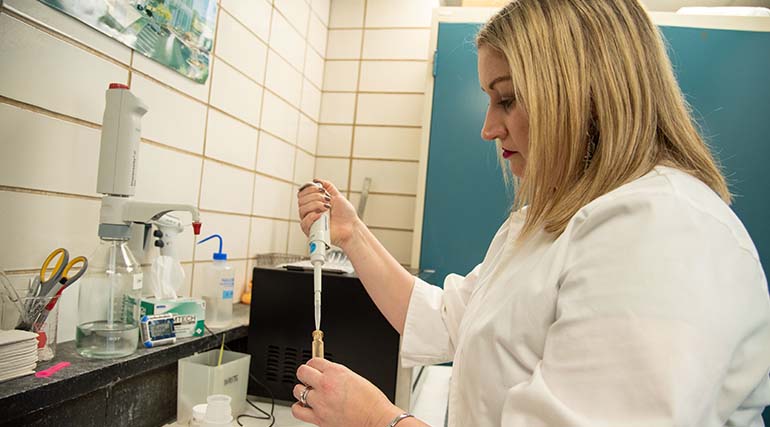 Person in white coat piping liquid into vial in laboratory.