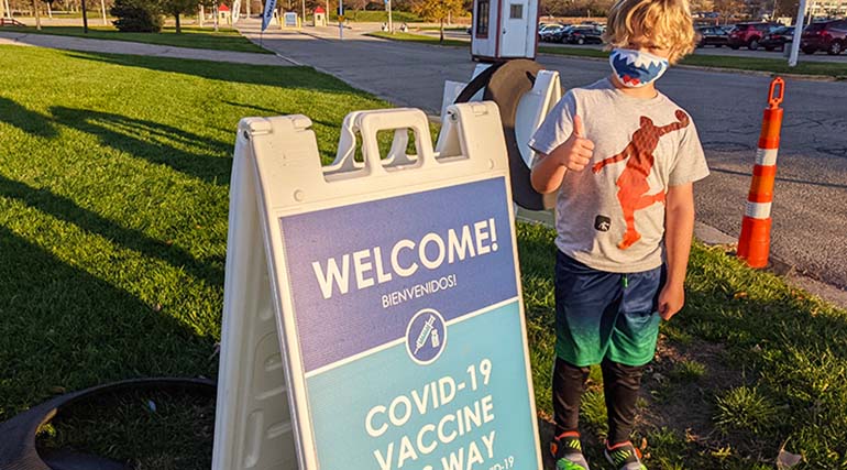 一名佩戴口罩的兒童在 COVID-19 疫苗接種標識牌旁邊豎起大拇指
