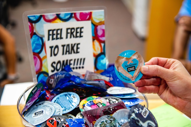A hand, holding a condom, next to a bowl of condoms and a sign that reads, "feel free to take some!"
