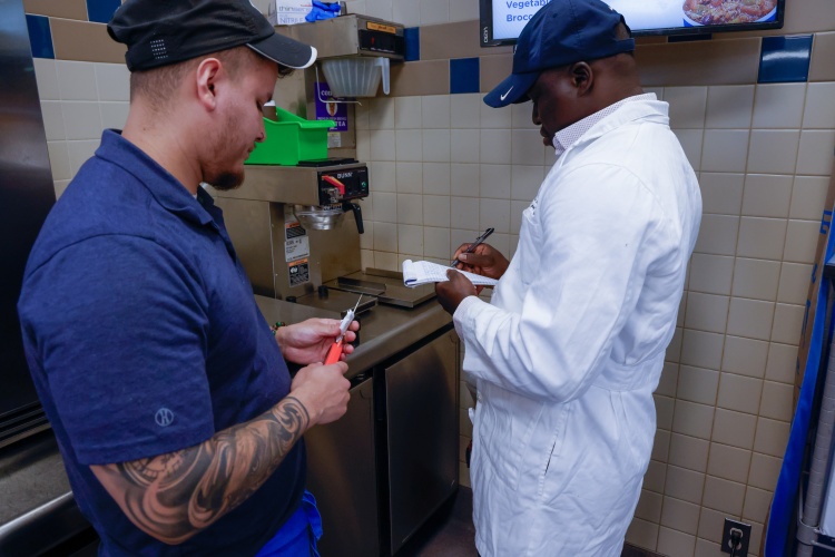 Public Health worker in white coat and blue hat holding note pad and pen with restaurant worker