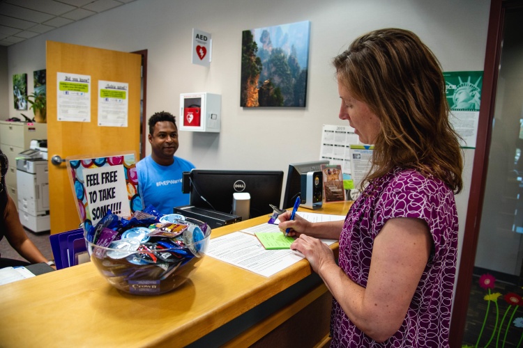 A person writing on paper standing at a counter, another person sitting behind the counter. 