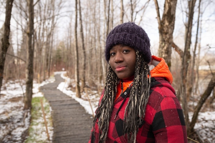 A smiling person wearing a winter coat and hat on a snowy trail with trees.