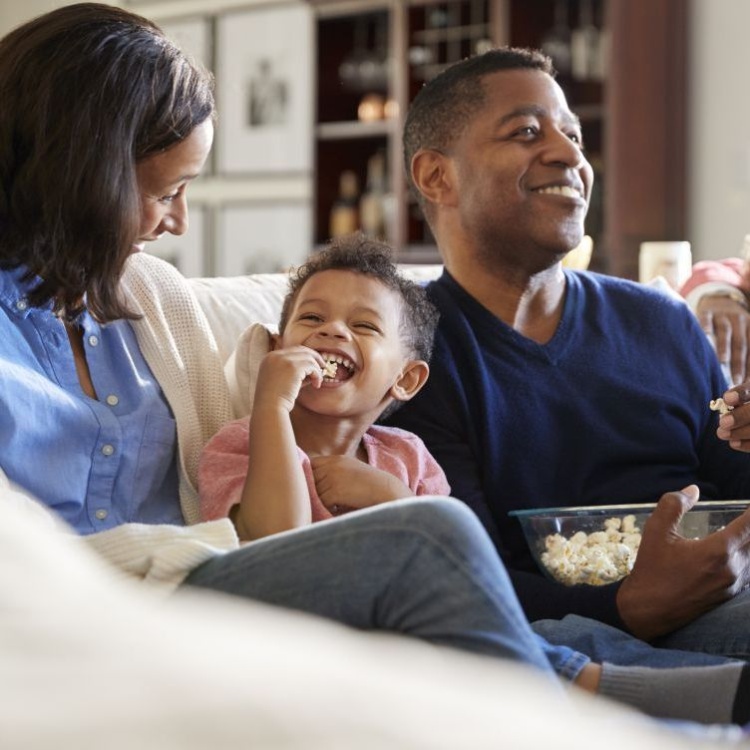 A family eats popcorn on the couch in their living room. They are smiling and happy.