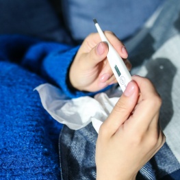 ill person holding a thermometer in bed with a tissue